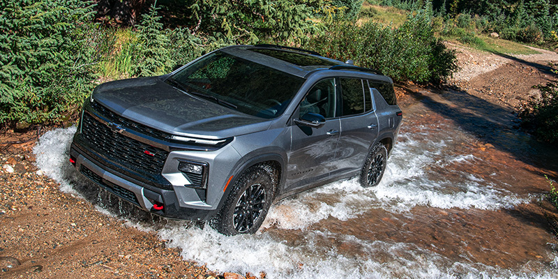 SUV driving through a shallow stream on an off-road trail, with water splashing around the wheels and a forested landscape in the background.