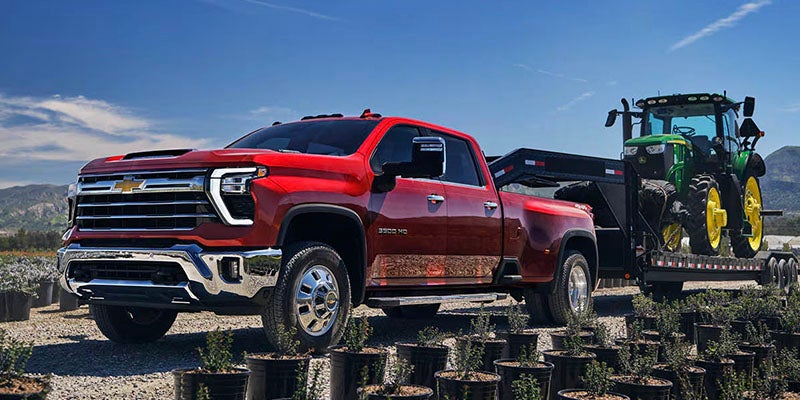 A red truck towing a green tractor on a flatbed trailer, set against a rural landscape.